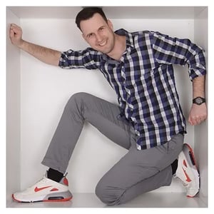 Timo Imbrogno Mitarbeiter von RIEDEL Networks kniet in einer weißen Box mit einem Arm an der Wand abgestützt. Trägt kariertes Hemd in Blau-Weiß, graue Hose und weiße Sneaker mit roten Details.